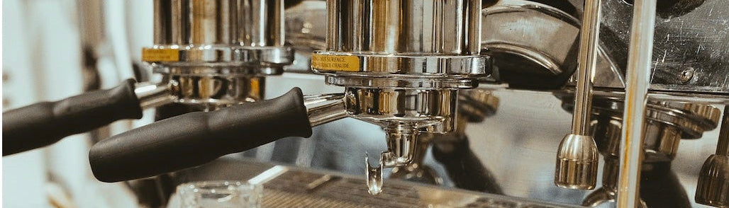 A close-up photograph of a professional espresso machine with two metal portafilters and small clear glass cups on the drip tray, shown within a white document or app interface.