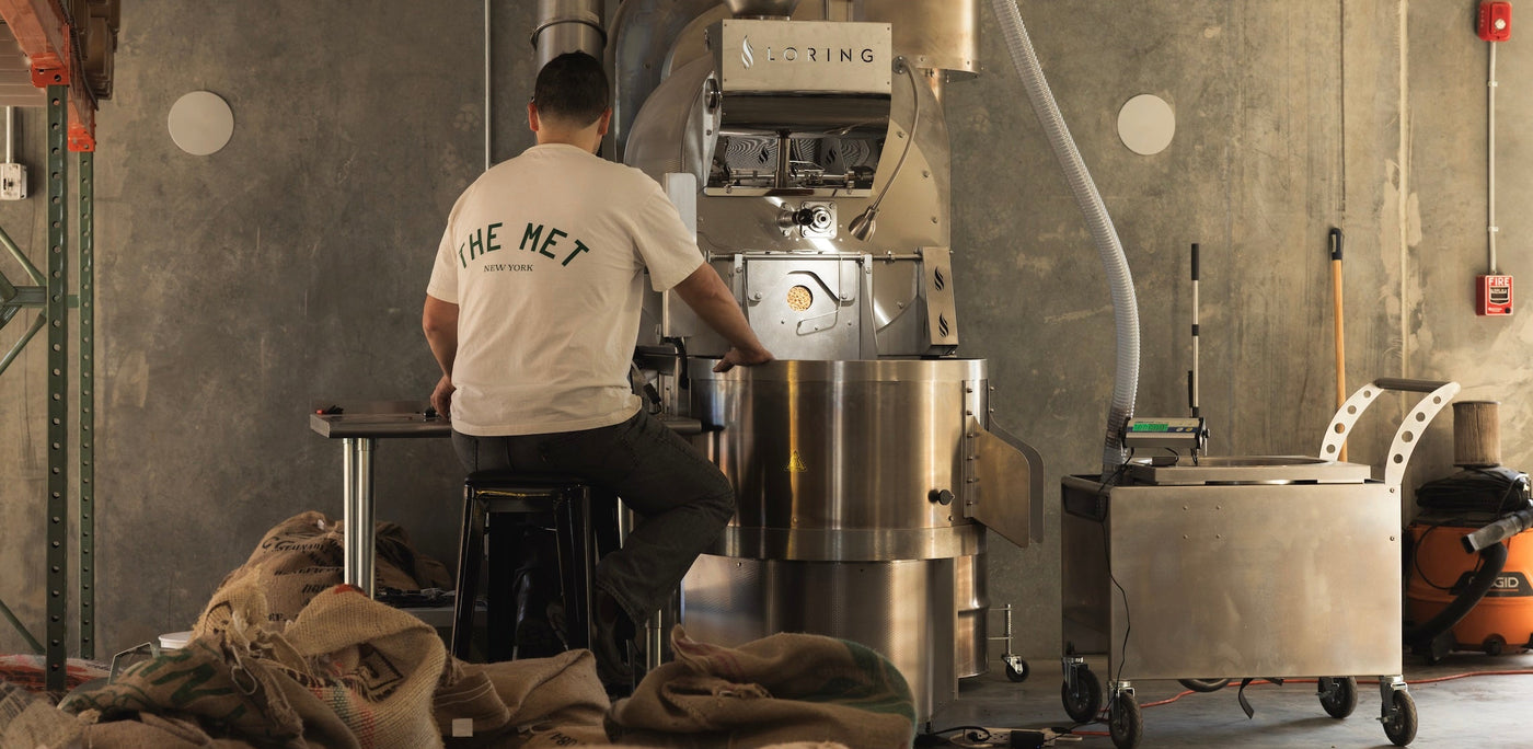 A man wearing a white t-shirt with 'THE MET' printed on the back sits on a stool in front of a large stainless steel coffee roaster, Loring, inside a warehouse roaster setting.